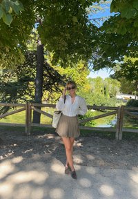 Λευκό πουκάμισο με μακριά μανίκια και κάθετες ρίγες, γκρι πλισέ φούστα, γκρι παπούτσια και μια ανοιχτόχρωμη τσάντα, σε φόντο ενός πράσινου πάρκου.