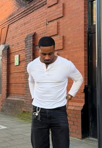 Homme en chemise blanche à manches longues et jean noir debout sur le trottoir ajustant son pantalon, avec des clés accrochées à la passants de ceinture près d'un bâtiment en brique.