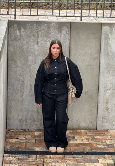 Mujer de cabello largo y castaño, vestida con chaqueta negra abotonada y pantalones, zapatos beige, y llevando un bolso beige al hombro, de pie frente a una pared de hormigón.
