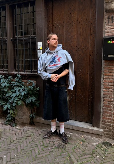 Hombre con el cabello recogido, vestido con ropa sobredimensionada en capas, de pie en una calle adoquinada frente a una puerta de madera con plantas cerca.