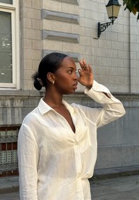 Camicia bianca a righe con vestibilità ampia e tasca, indossata con i capelli raccolti. Completata da orecchini a cerchio dorati.