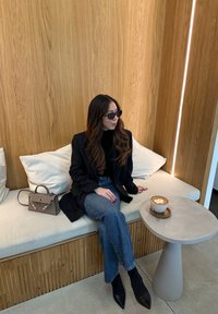 Μαύρο σακάκι πάνω από μια εφαρμοστή μαύρη μπλούζα, μπλε ψηλόμεσα τζιν, μαύρες μυτερές γόβες, καθισμένο σε έναν ανοιχτό καναπέ με ένα στρογγυλό γκρι κομοδίνο και καφέ.