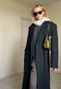 Cappotto nero doppiopetto in tessuto strutturato, maglione grigio lavorato a maglia, pantaloni beige e borsa verde. Accessoriato con occhiali da sole neri.