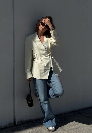 Mujer con blazer beige con cinturón, vaqueros azules, gafas de sol, sosteniendo un bolso negro, apoyada en la pared con una pierna levantada.