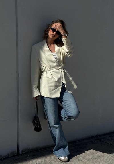 Mujer con blazer beige con cinturón, vaqueros azules, gafas de sol, sosteniendo un bolso negro, apoyada en la pared con una pierna levantada.