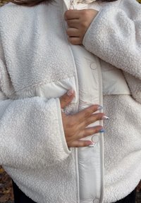 Jachetă albă pufoasă cu un panou din material neted contrastant, închideri cu capse și mâneci largi. Prezintă o mână sprijinită pe partea din față.