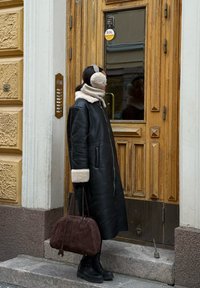 Manteau en cuir noir avec poignets en shearling beige, sac à main en daim marron, et bottes de combat noires, debout près d'une porte en bois avec un panneau en verre.