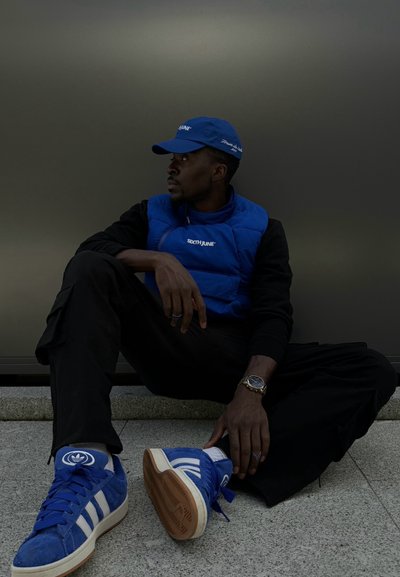 Chaleco acolchado azul y gorra, camiseta de manga larga negra, pantalones negros. Zapatillas de ante azules con franjas blancas. Fondo texturizado gris.