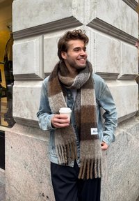 Écharpe : motif écossais marron et gris avec des bords à franges, fabriquée en matériau doux. Portée avec une veste en denim bleu clair et tenant une tasse de café.