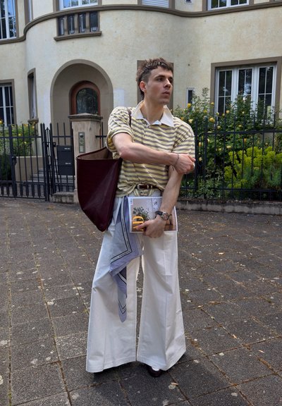 Joven con una camisa polo a rayas y pantalones blancos de pierna ancha está de pie en un camino pavimentado sosteniendo una revista y una bolsa tote granate.