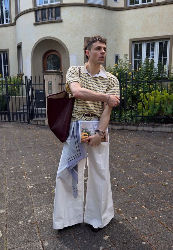 Ein junger Mann trägt ein gestreiftes Polohemd und weiße Hose mit weitem Bein. Er steht auf einem gepflasterten Weg und hält ein Magazin sowie eine dunkelrote Tragetasche.