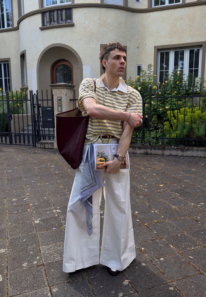Ein junger Mann, der ein gestreiftes Poloshirt und weiße Hose mit weitem Bein trägt, steht auf einem gepflasterten Weg und hält ein Magazin sowie eine weinrote Tragetasche.