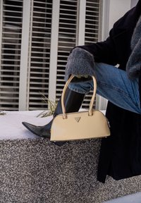 Άτομο που φοράει γκρι γάντια και μαύρες μπότες κρατάει μπεζ τσάντα σχεδιαστή ενώ στηρίζεται με το πόδι σε πεζούλι από πέτρα σε εξωτερικό χώρο.