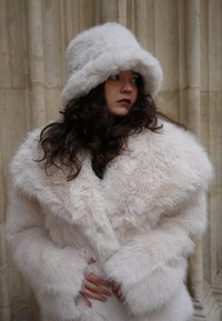 Plašč iz umetnega krzna v nežni svetlo roza barvi z velikim ovratnikom in širokimi rokavi; ujemajoča se puhasta bela kapa; teksturirana površina dodaja globino.