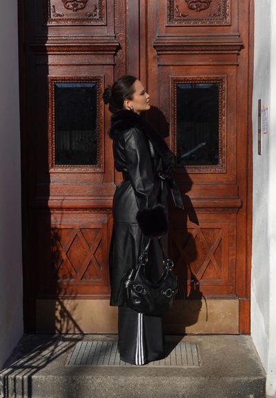 Mujer con abrigo de cuero negro con borde de piel y bolso negro, de pie frente a una gran puerta de madera adornada bajo la luz del sol.