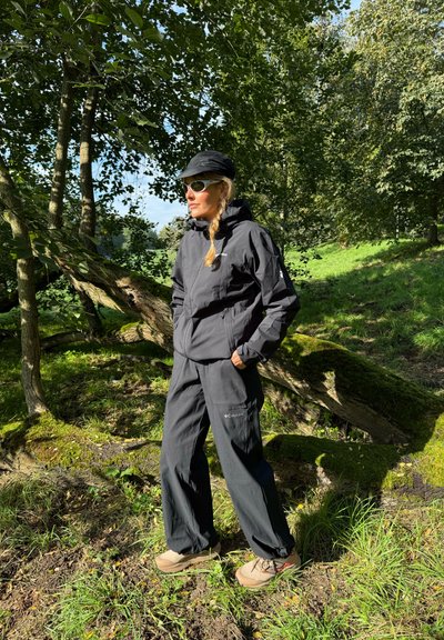 Chaqueta impermeable negra y pantalones holgados, ambos con un acabado texturizado, combinados con botas de senderismo beige. Gafas de sol y una gorra completan el look.