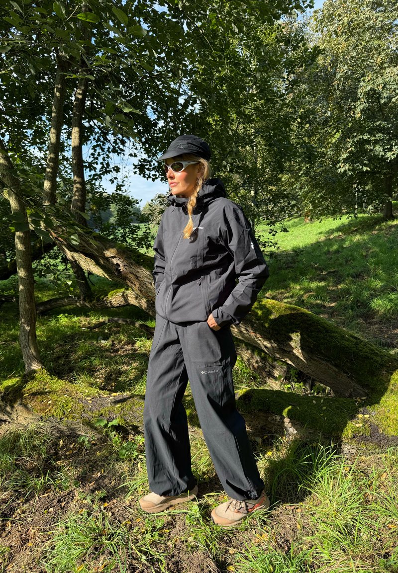 Fekete vízálló dzseki és bő szabású nadrág, mindkettő texturált felülettel, bézs túrabakancsokkal párosítva. A napszemüveg és sapka teszi teljessé a megjelenést.