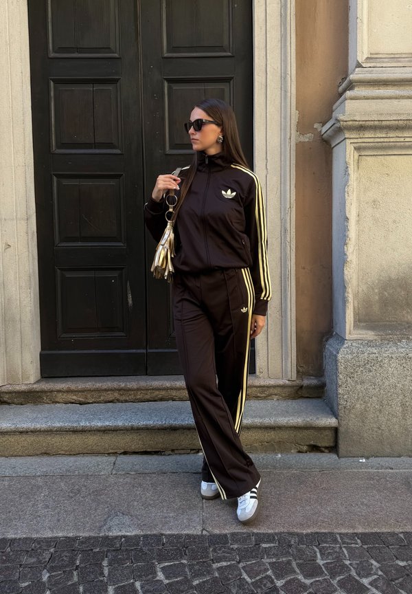 Frau trägt schwarzen Adidas-Trainingsanzug mit gelben Streifen, weiße Turnschuhe, Sonnenbrille und hält eine metallisch goldene Handtasche, steht an einer Tür.