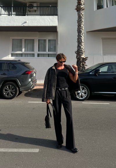 Persona con una camiseta sin mangas negra y pantalones anchos negros, sosteniendo una bolsa. Lleva encima una chaqueta de estampado de leopardo, en un entorno urbano.