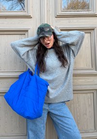 Pull gris oversize, sac cabas matelassé bleu, casquette de baseball camouflage, lunettes transparentes, debout contre un panneau en bois. Matériaux texturés.