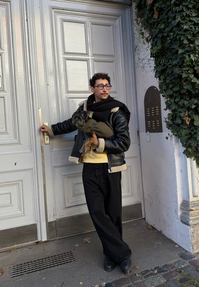 Hombre con chaqueta de cuero negra y bufanda sostiene un perro pequeño mientras abre una gran puerta de madera junto a una pared cubierta de hiedra.