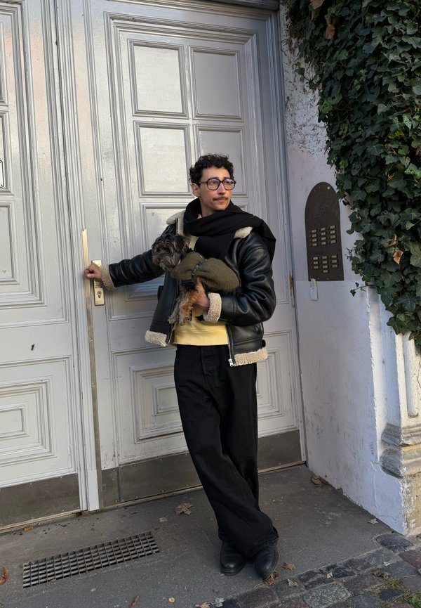 Ein Mann trägt eine schwarze Lederjacke und einen Schal, hält einen kleinen Hund und öffnet eine große Holztür neben einer mit Efeu bewachsenen Wand.