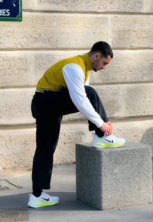 Hombre con chaleco amarillo y camisa blanca atándose un zapato Nike blanco con verde neón sobre un bloque de piedra al aire libre junto a una pared texturizada.