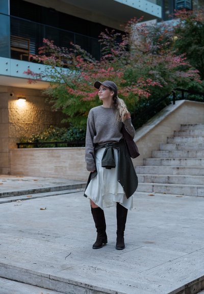Suéter gris, falda blanca con diseño floral, camisa a cuadros verde atada en la cintura, botas altas marrones, gorra, y un fondo de escalones de piedra y vegetación.