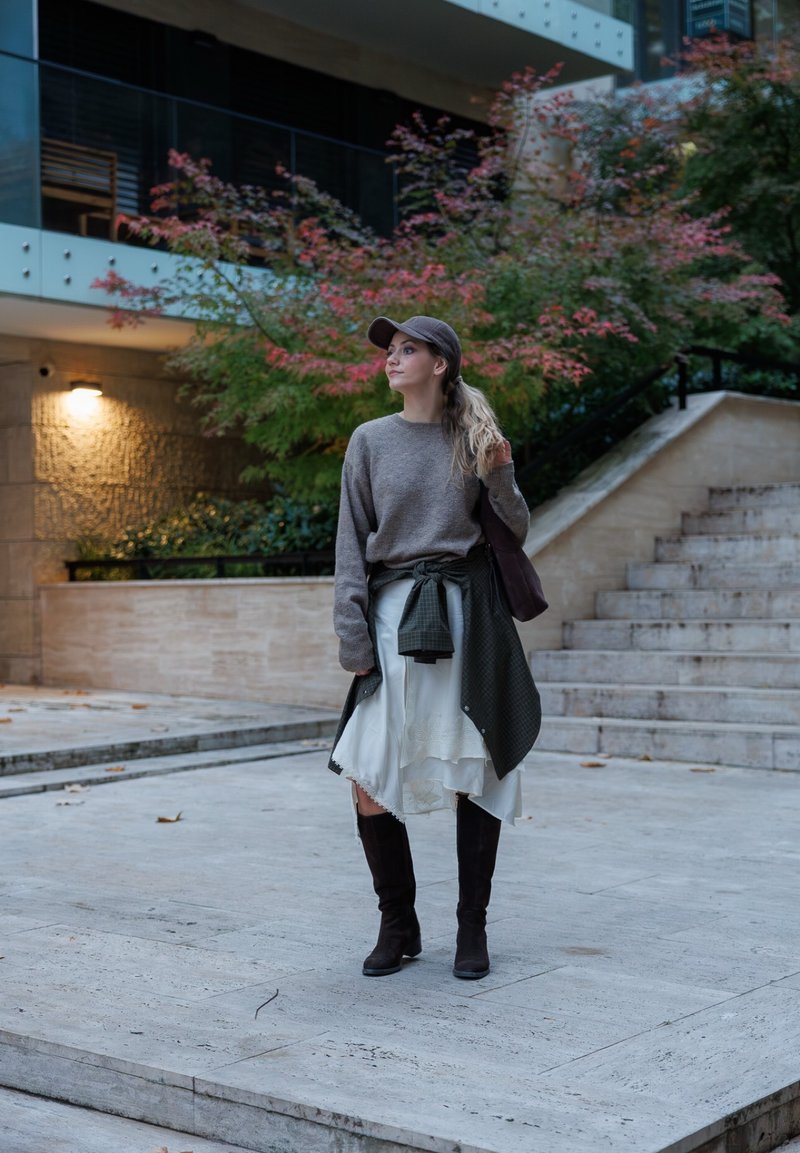 Sivi pulover, bijela suknja s cvjetnim uzorkom, zelena karirana košulja vezana oko struka, visoke smeđe čizme, kapa, a u pozadini su kameni stepenice i zelenilo.