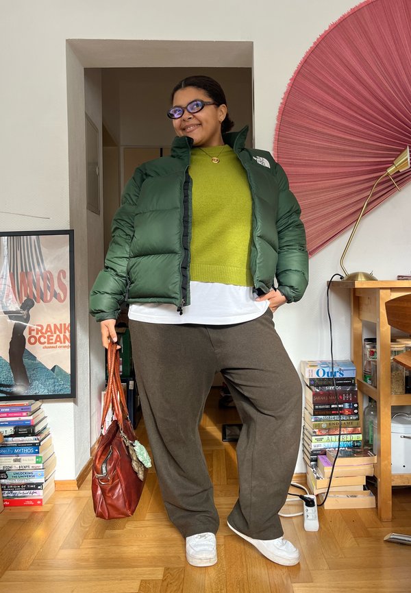 Personne portant une doudoune verte, un pull vert citron, un pantalon foncé, des baskets blanches, tenant un sac en cuir marron, debout à l'intérieur près de livres et d'œuvres d'art.