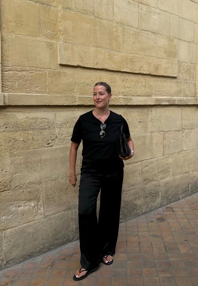 Top negro de manga corta y pantalones anchos negros, complementados con gafas de sol y un bolso de mano tejido negro, frente a una pared beige texturizada.