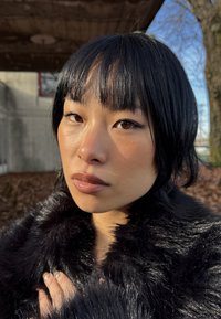 Jeune femme aux cheveux courts noirs, portant un manteau en fausse fourrure noire, debout en plein air sous le soleil avec un mur en béton et un arbre en arrière-plan.