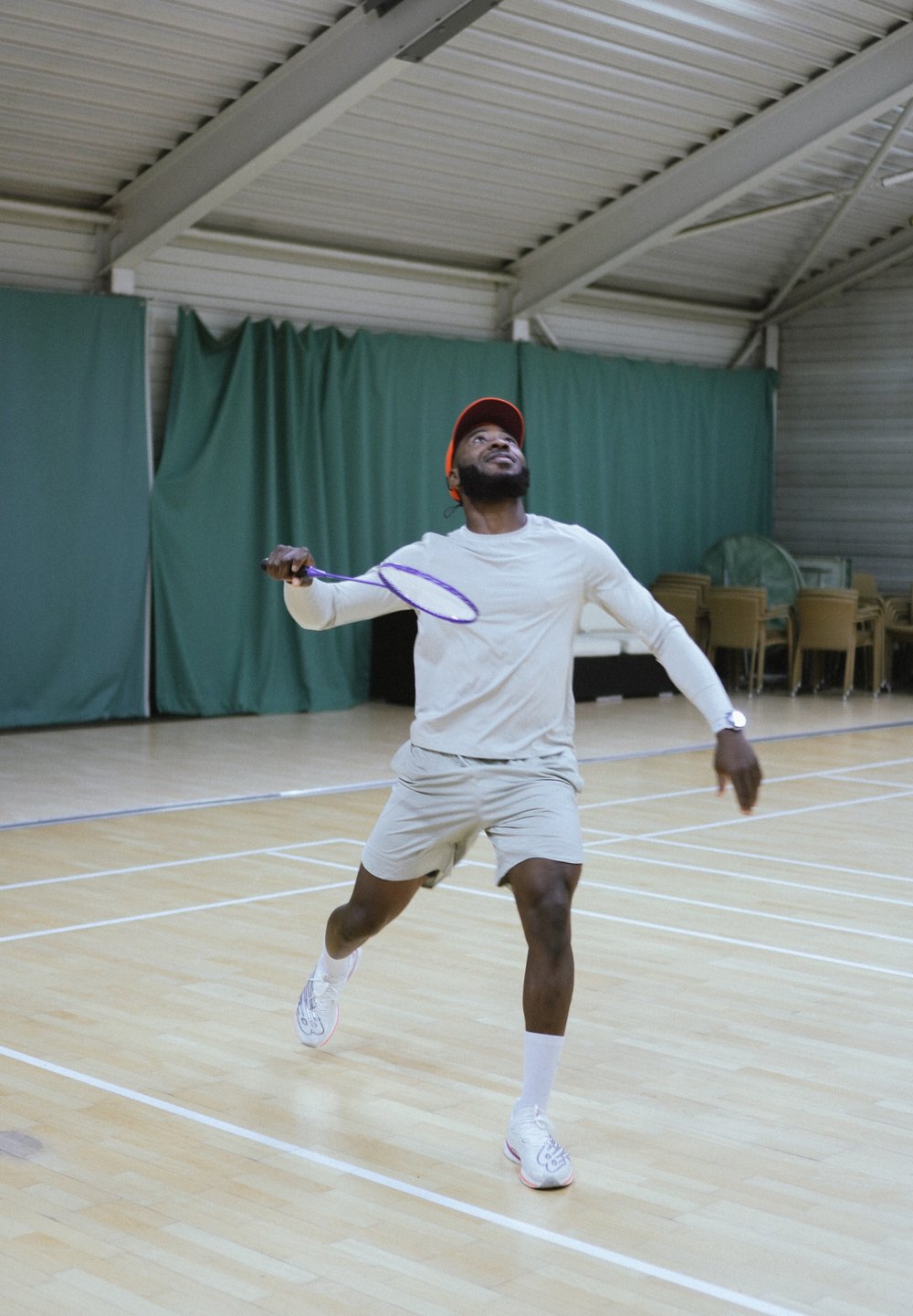Un homme vêtu d'une tenue de sport gris clair et d'une casquette orange se prépare à frapper un volant avec une raquette de badminton violette en intérieur.