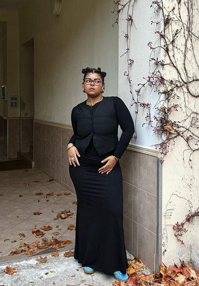 Falda larga negra ajustada y top negro de manga larga con chaleco abotonado. Calzado azul. El fondo presenta una pared de azulejos beige y hiedra.