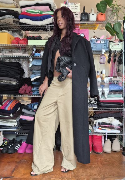 Abrigo largo negro, camiseta corta negra, pantalones beiges de pierna ancha y bolso de mano negro tejido. El fondo presenta ropa y accesorios apilados ordenadamente.