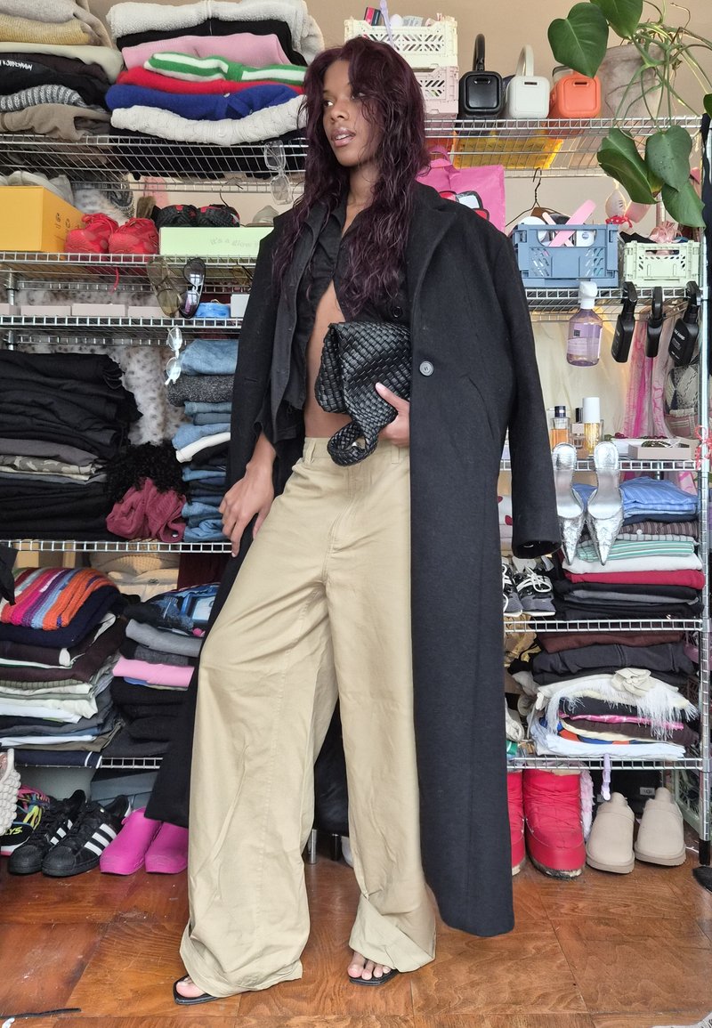 Black long coat, cropped black shirt, beige wide-leg trousers, and woven black clutch. Background features neatly stacked clothing and accessories.