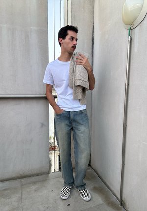 Hombre con camiseta blanca, vaqueros azules y zapatos slip-on a cuadros, de pie en el balcón, sosteniendo un suéter de punto beige sobre su hombro.