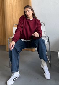 Felpa bordeaux a maniche lunghe, abbinata a un paio di jeans in denim blu larghi e sneakers bianche con strisce nere, seduto su una sedia.