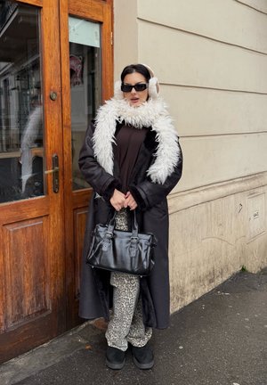 Abrigo largo negro con cuello de piel, camiseta marrón, pantalones con estampado de leopardo, bolso negro con correas y orejeras. Cabello oscuro y gafas de sol.