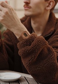 Braune Fleecejacke mit einer flauschigen Textur. Eine bronzefarbene Digitaluhr mit quadratischem Ziffernblatt ist sichtbar an einer Hand, die eine Tasse hält, neben einem weißen Teller.