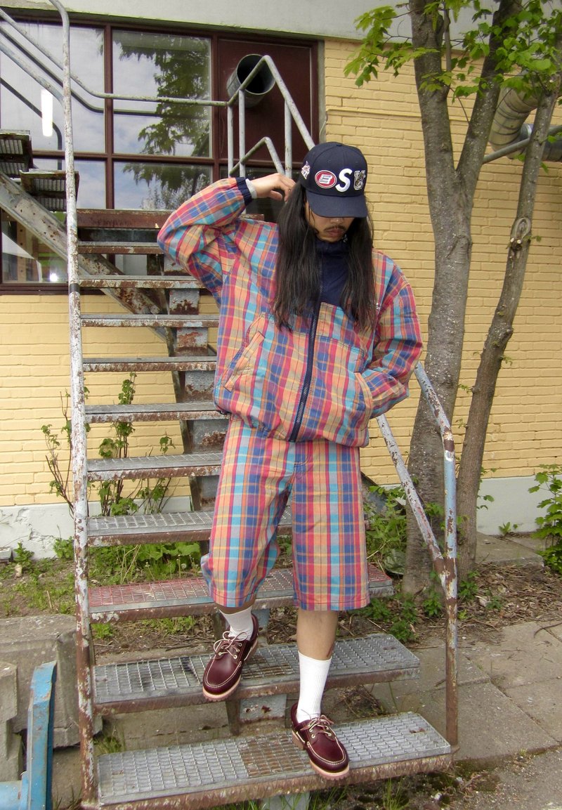 Person iført matchende farverig ternet jakke og shorts med bordeaux sko, hvide sokker og en sort kasket, stående på udendørs metaltrapper.
