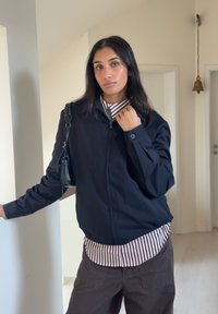 Veste noire zippée au design ajusté, associée à une chemise rayée blanche et rouge, portée avec un pantalon marron foncé. Sac à main noir en accessoire.