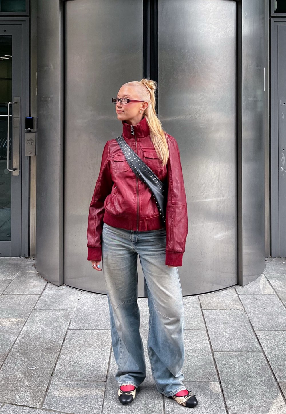 Giacca di pelle rossa con colletto alto e tasche a toppa, abbinata a jeans baggy blu chiaro e scarpe bianche e nere. Lo sfondo presenta porte metalliche.