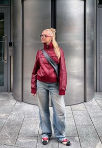 Giacca di pelle rossa con colletto alto e tasche a toppa, abbinata a jeans baggy blu chiaro e scarpe bianche e nere. Lo sfondo presenta porte metalliche.