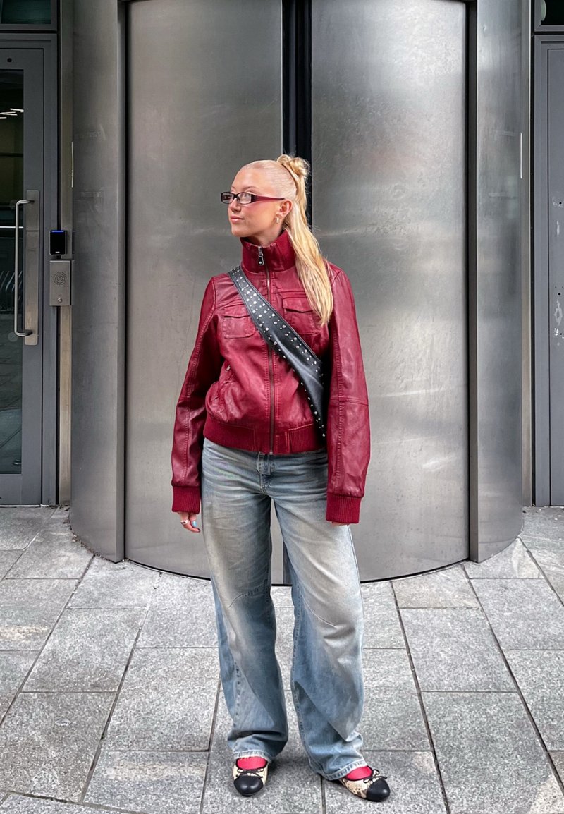 Piros bőrdzseki magas gallérral és foltzsákokkal, világoskék, laza farmernadrággal és fekete-fehér cipőkkel párosítva. A háttér fém ajtókkal van díszítve.