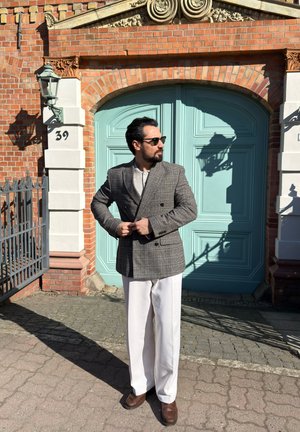 Hombre con gafas de sol, blazer de cuadros gris, pantalones blancos y zapatos marrones de pie frente a una puerta arqueada color verde azulado con ladrillos y moldura blanca.