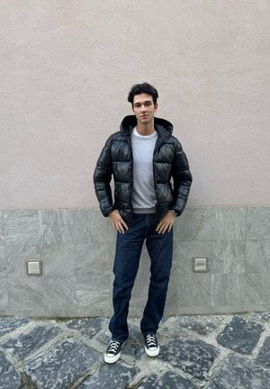 Chaqueta de plumas negra con un acabado brillante, suéter gris, jeans azul oscuro y zapatillas negras, exhibidos contra una pared texturizada clara.