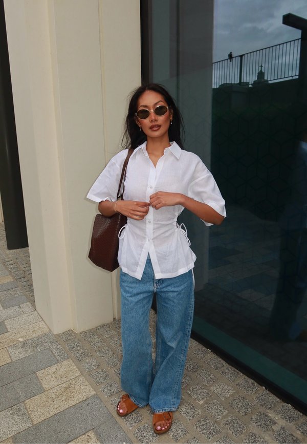 Donna che indossa una camicia bianca abbottonata, jeans blu, sandali marroni e occhiali da sole, in piedi all'aperto vicino a una parete di vetro e a un edificio beige.