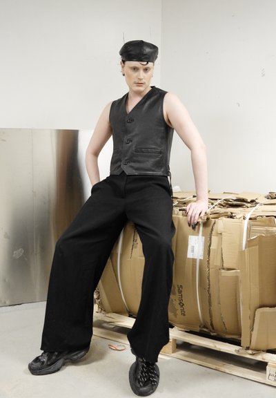 Persona con chaleco de cuero negro, pantalones anchos negros, zapatillas negras y boina de cuero negra, sentada sobre cajas de cartón apiladas en un almacén.