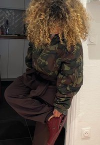 Casaco bomber de camuflagem verde com textura acolchoada, combinado com cal�ças largas escuras e ténis Nike bordô, em ambiente interior.